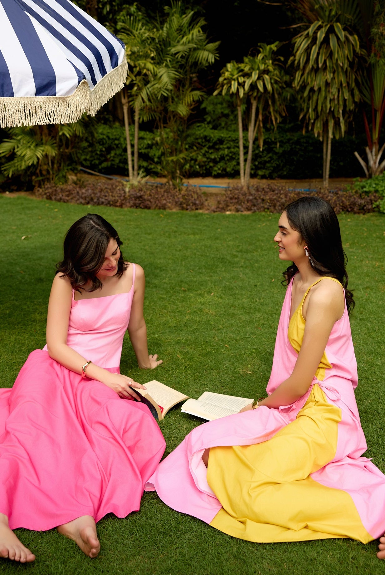 Two women in colorful dresses sitting on grass under a striped umbrella.