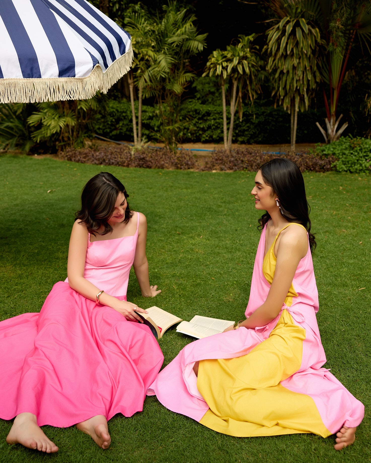 Two women in colorful dresses sitting on grass under a striped umbrella.