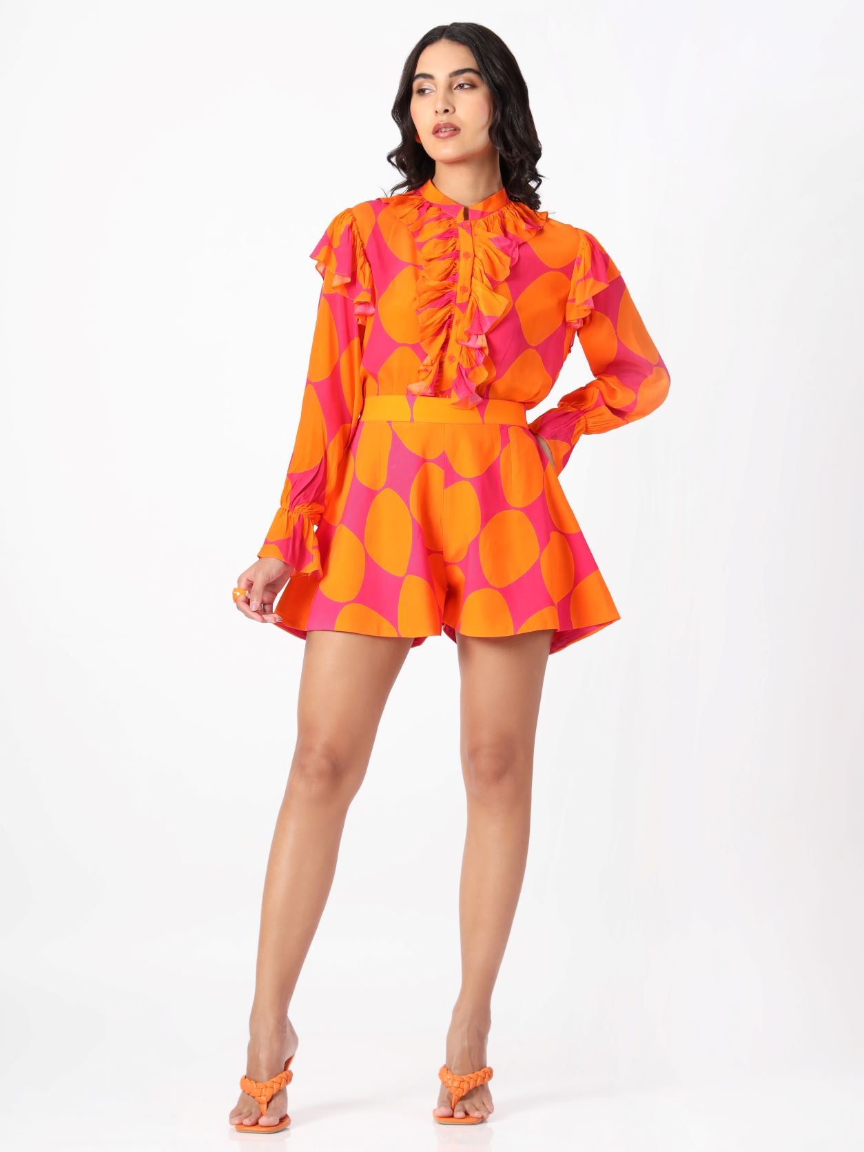 A model wearing a long sleeve shirt with a ruffled neckline and ruffles on the sleeves, paired with matching shorts, in a vibrant pink and orange geometric pattern.